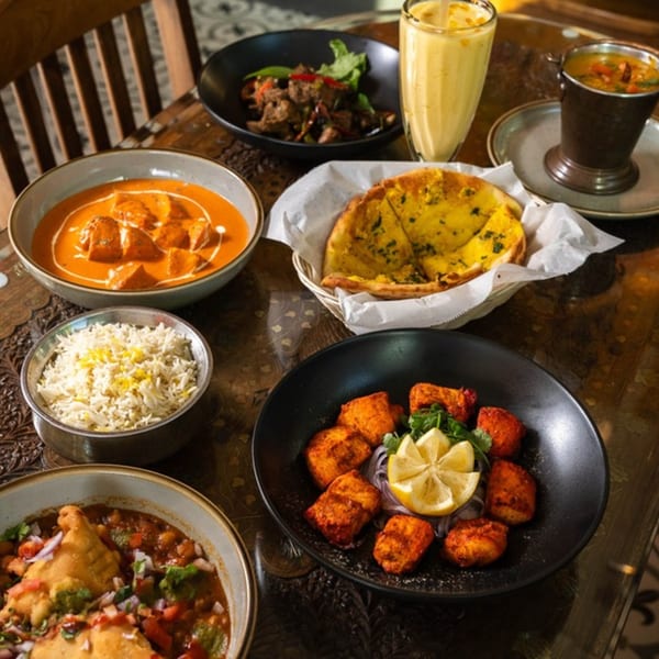 A table laden with various Indian dishes, including butter chicken, rice, naan, samosa chaat, and mango lassi.