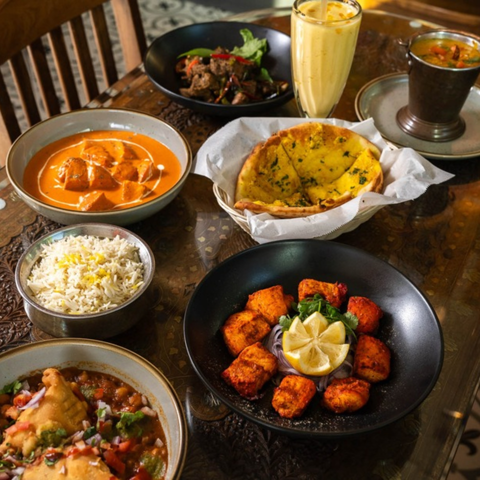 A table laden with various Indian dishes, including butter chicken, rice, naan, samosa chaat, and mango lassi.