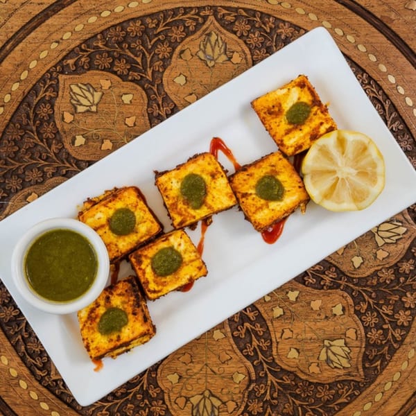 Indian paneer tikka with green chutney and lemon on a white plate, on a wooden table.