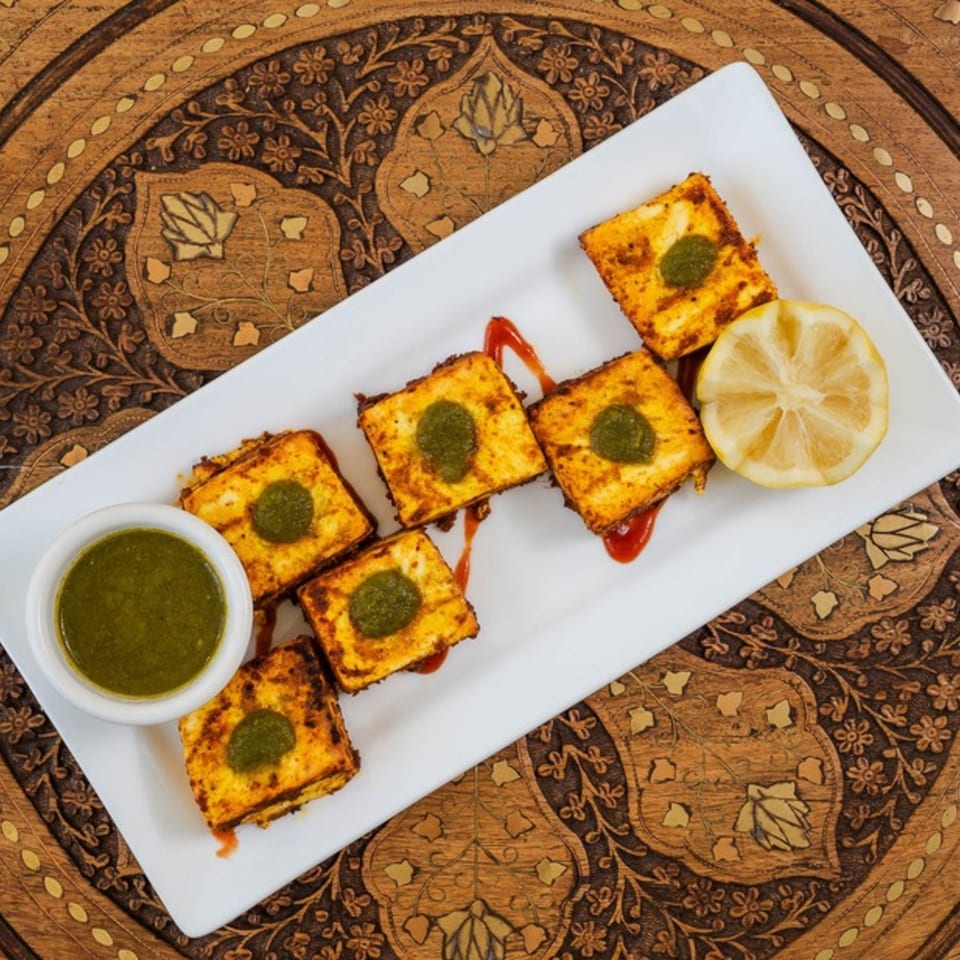 Indian paneer tikka with green chutney and lemon on a white plate, on a wooden table.