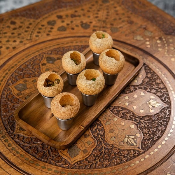 Six pani puri in metal cups on a wooden tray, on a carved wooden table.