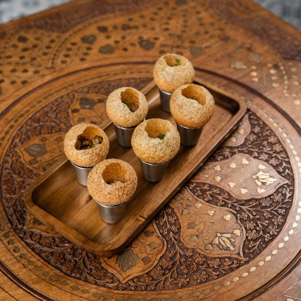 Six pani puri in metal cups on a wooden tray, on a carved wooden table.