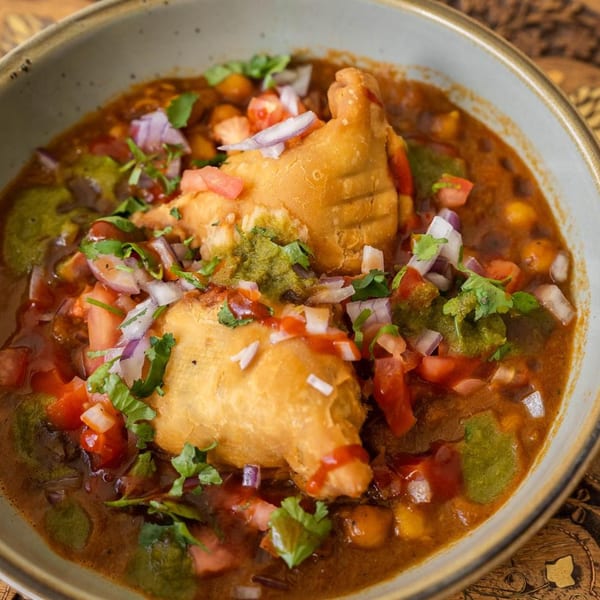 Two golden-brown samosas submerged in a rich, spicy chickpea curry, garnished with fresh cilantro, diced tomatoes, and red onions.