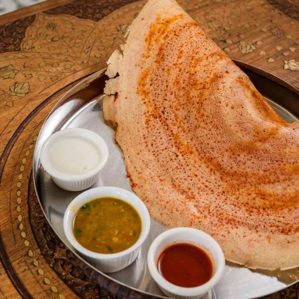 A crispy dosa with three small bowls of sauces on a silver plate, on a wooden table.